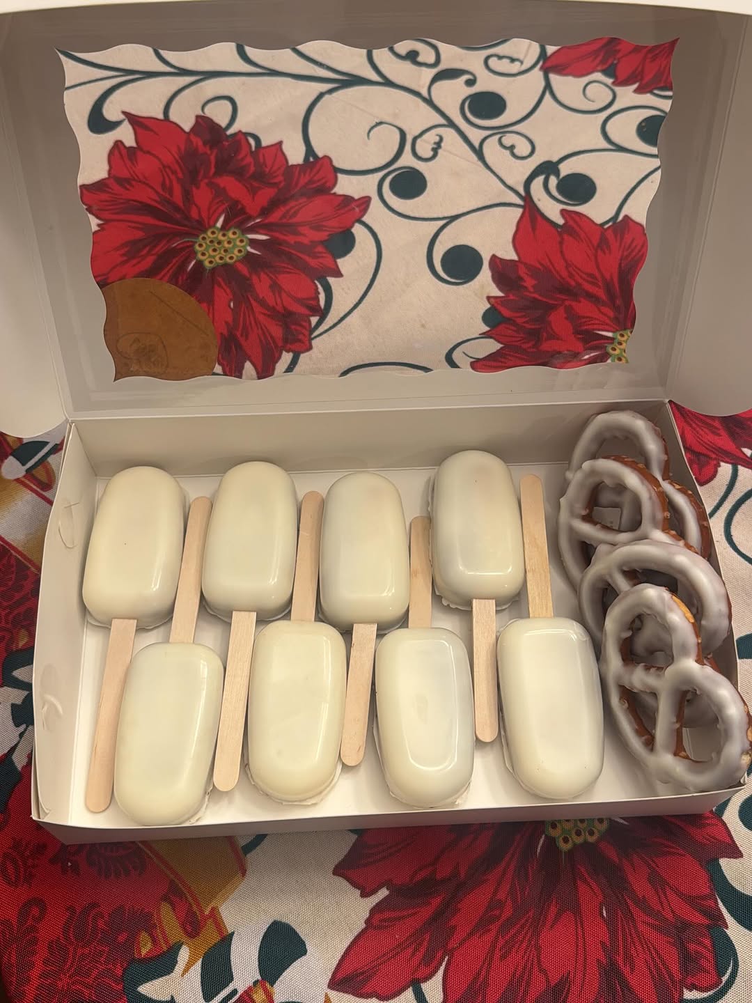 Elegant all-white chocolate cakesicles and pretzels in a white bakery box, with a festive red poinsettia holiday tablecloth behind.