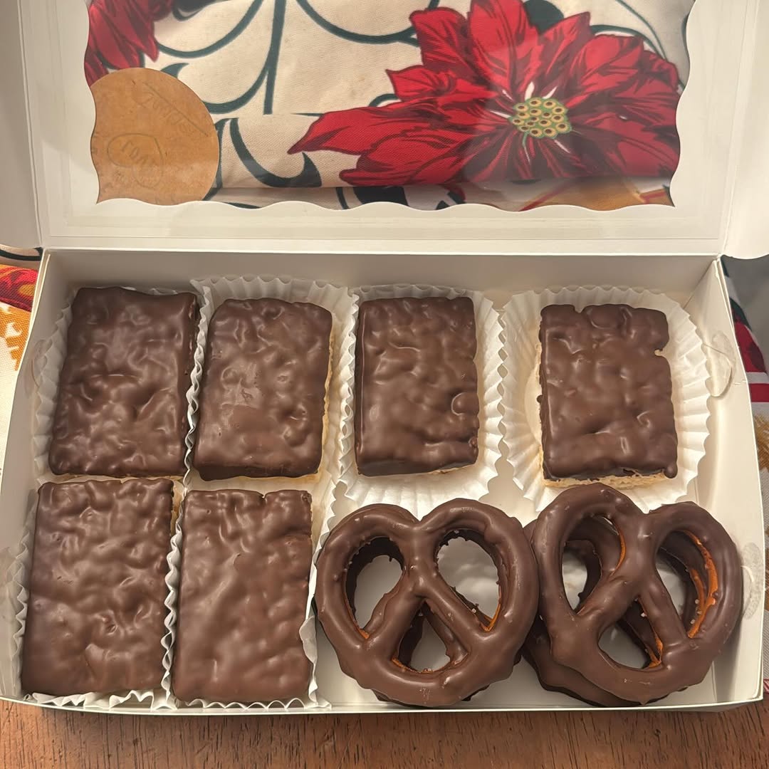 Holiday gift box of six chocolate-covered Rice Krispie bars and two large pretzels in smooth milk chocolate, with a festive poinsettia backdrop.