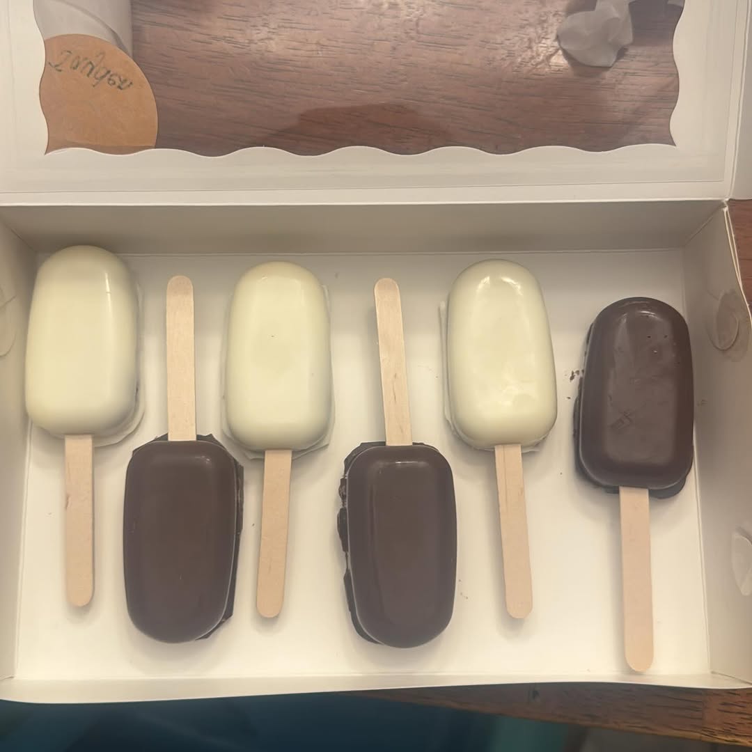 Six cakesicles alternating between white ivory and dark chocolate coatings, arranged in a single clean row inside a white bakery box.