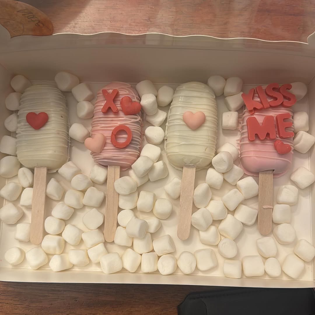 Four Valentine's Day cakesicles decorated with red fondant hearts, XO letters, and KISS ME text, standing upright in a bed of marshmallows.