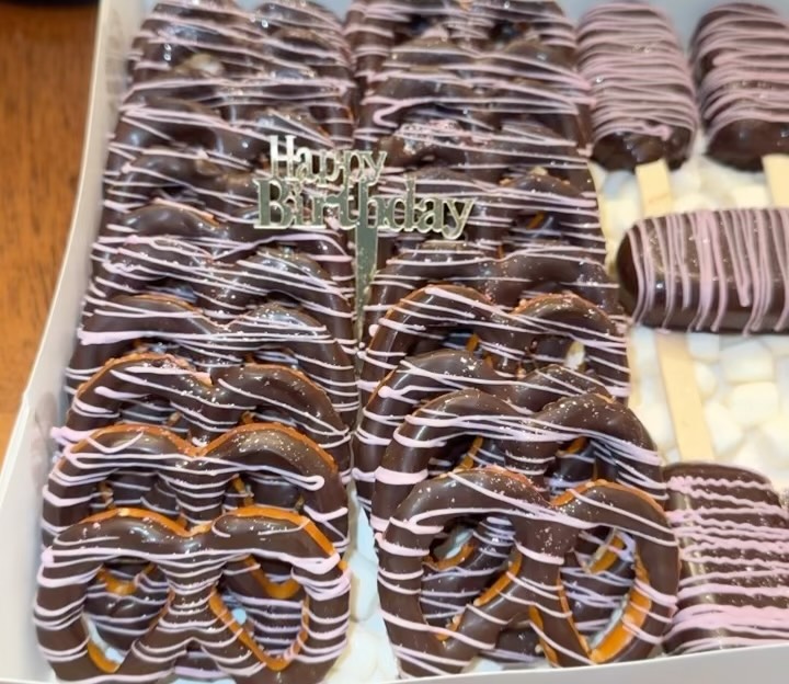Close-up of dark chocolate pretzels with pink and white wave drizzle and a gold metallic Happy Birthday script topper pick.