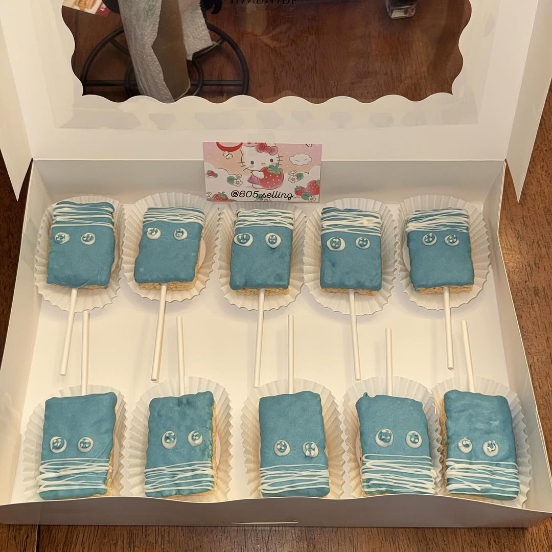 Ten teal blue chocolate-dipped Rice Krispie treat pops on sticks with white drizzle stripes and button candy accents in a white bakery box.