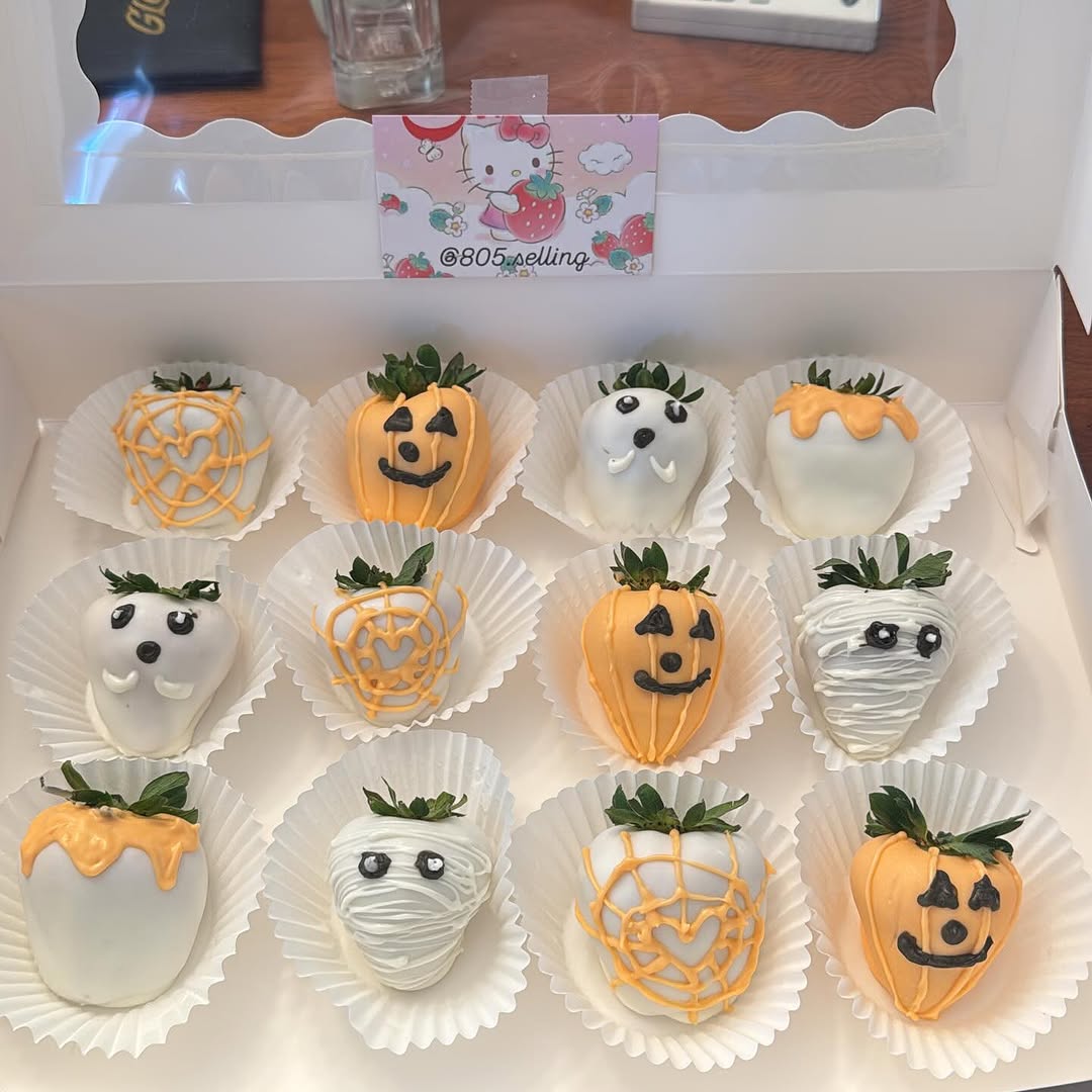 A dozen Halloween-themed chocolate-covered strawberries featuring ghost, jack-o-lantern, spiderweb, and mummy designs in white and orange coatings.