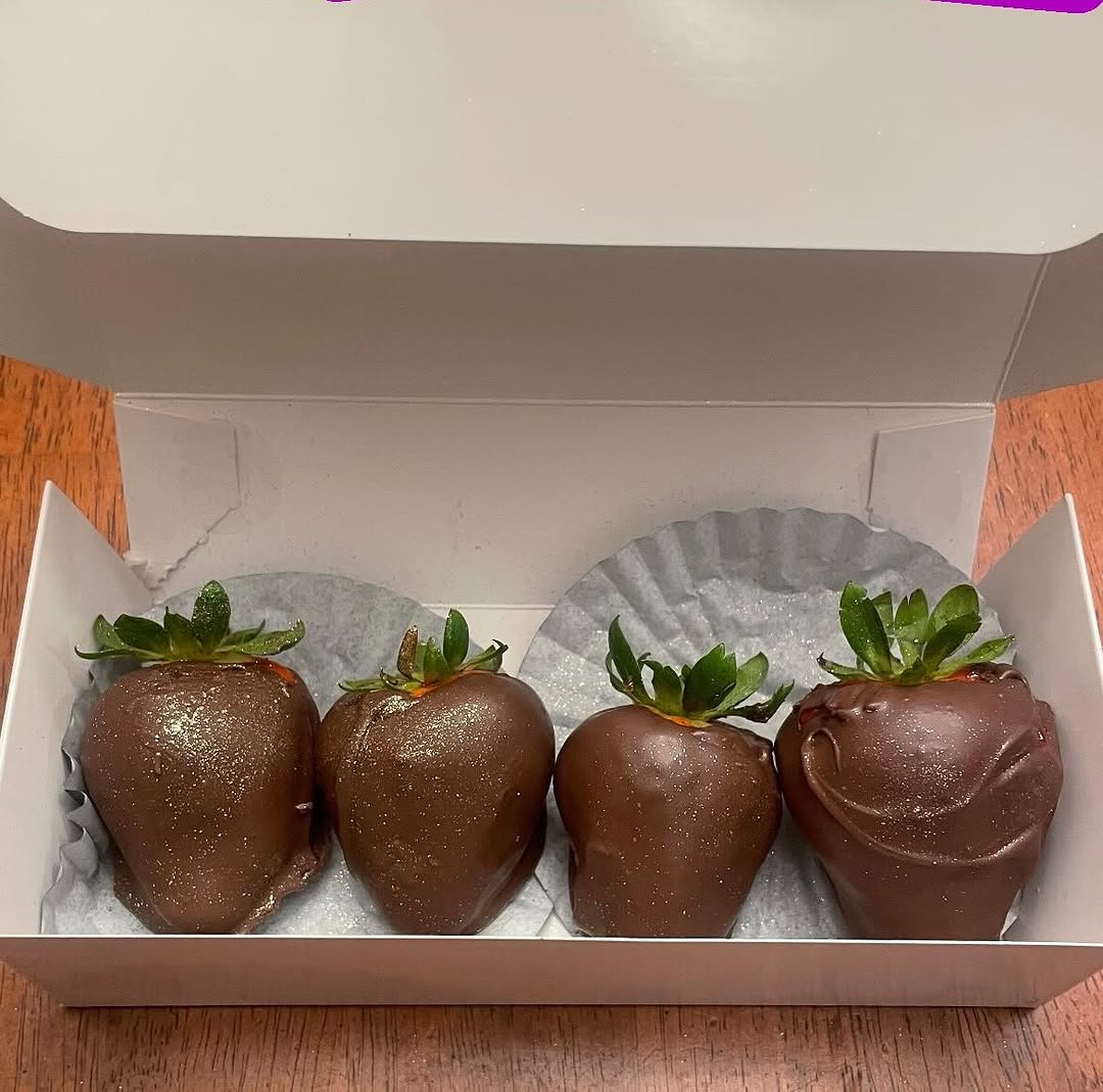 Four large milk chocolate-covered strawberries dusted with gold edible glitter, lined up in a small white bakery box.