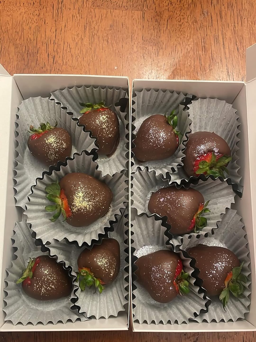 Eight dark chocolate-covered strawberries with gold shimmer dust arranged across two small white boxes, photographed from above.