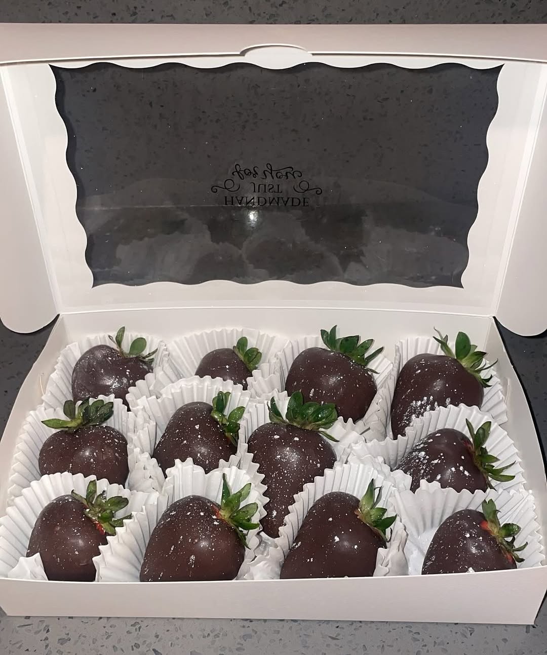 A dozen dark chocolate-covered strawberries with white shimmer dusting, arranged in a four-by-three grid in a white bakery box.